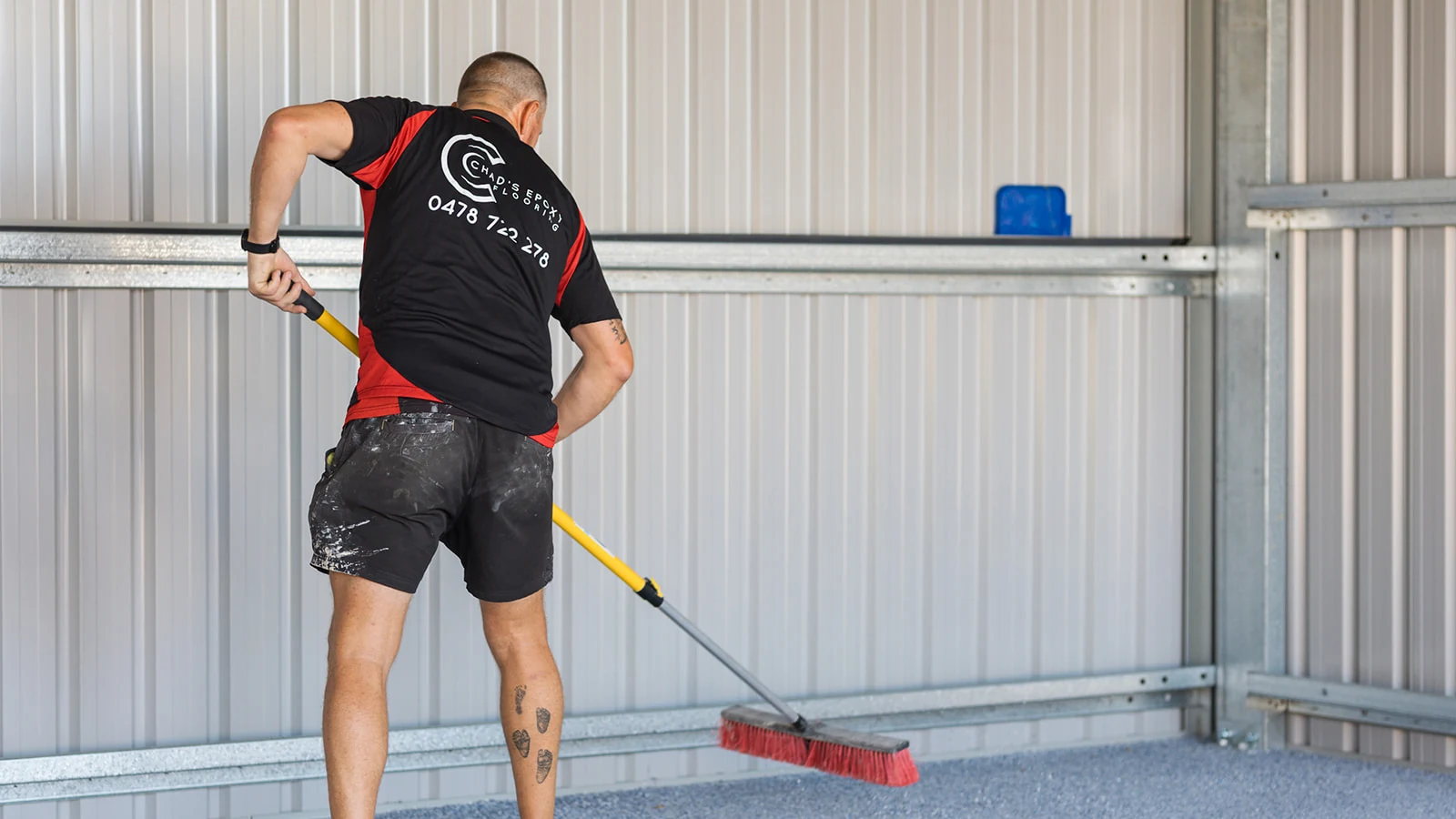 Epoxy installer sweeping excess flakes along the wall of a steel shed
